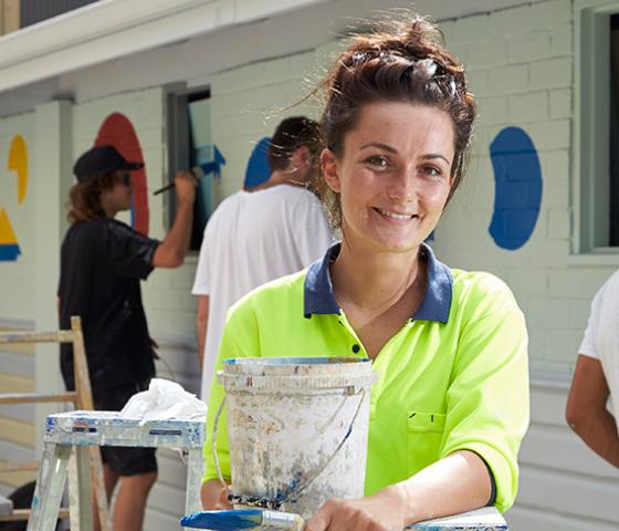 Image of a person wearing a hivis t-shirt, holding a bucket of paint. People in the background are painting a wall 